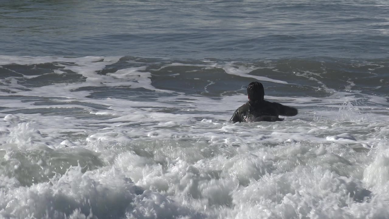 A dark-clad surfer paddles away from camera through choppy Huntington Beach water as a white wave rolls across the foreground.