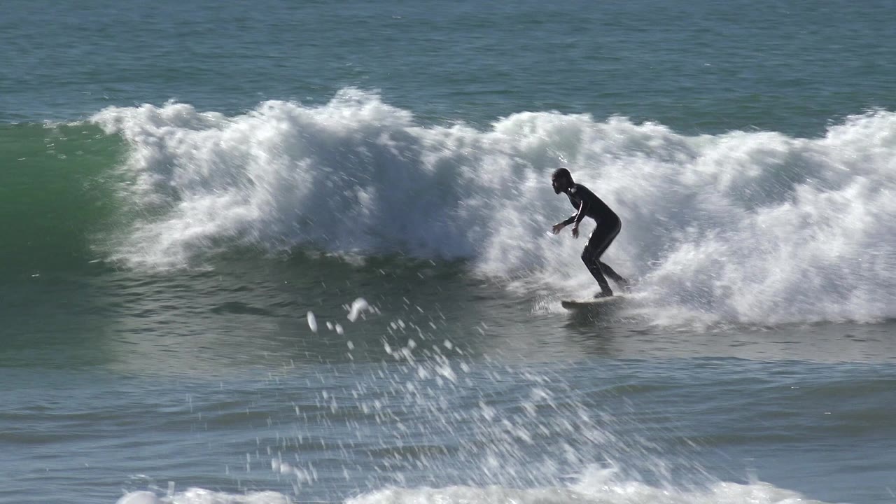 A surfer in a dark wetsuit rides across a breaking green-blue wave at Huntington Beach, California, with white spray behind the board.