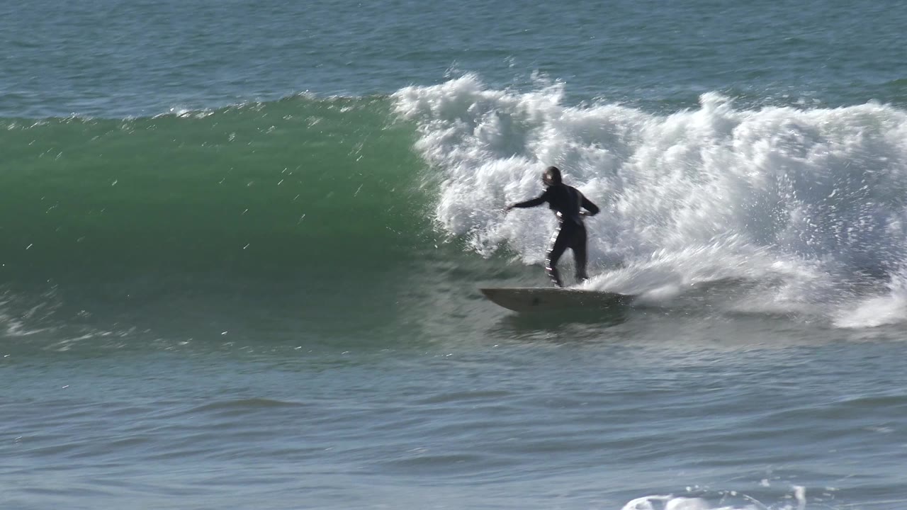 A distant surfer in a dark wetsuit rides near the right side of a breaking wave at Huntington Beach, California, with heavy foam trailing behind.