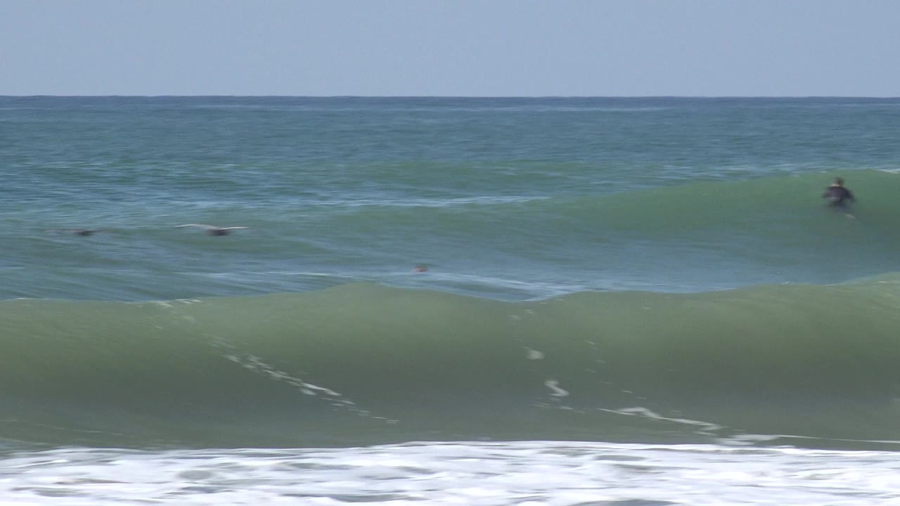 A wide Huntington Beach ocean view shows a green-blue swell rising behind foreground foam, with small distant surfers in the water and no close faces visible.