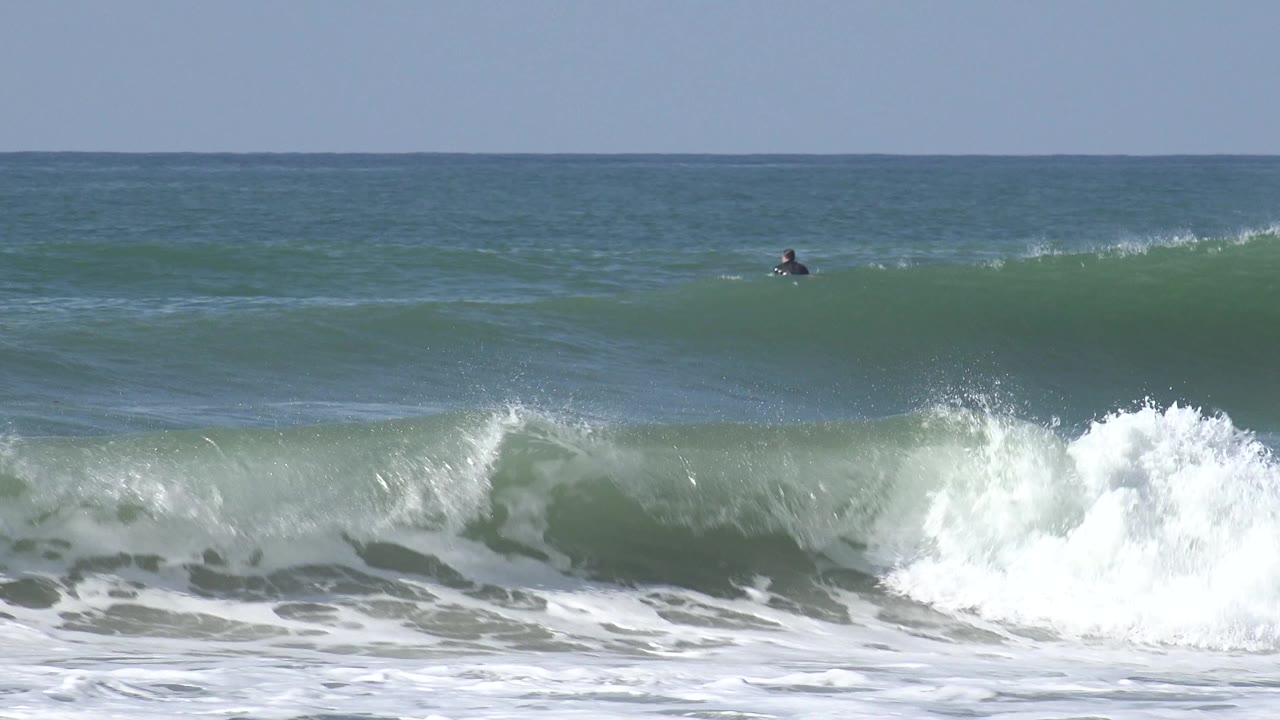 Whitewater breaks across the foreground at Huntington Beach as distant surfers sit farther out in the darker blue-green water under a clear horizon.