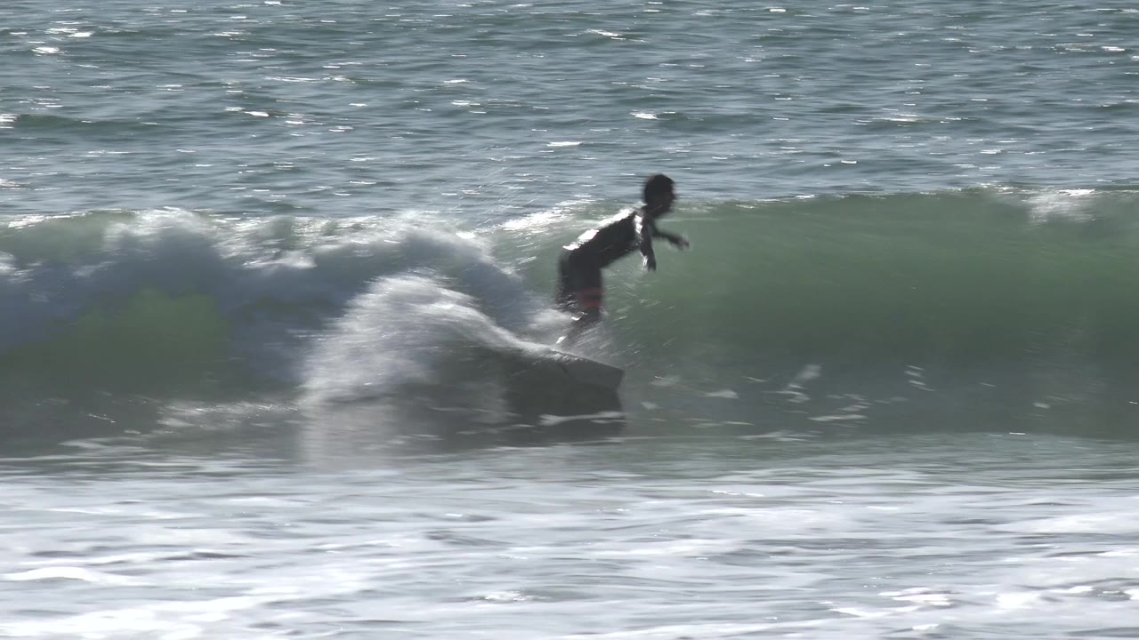 A young surfer stands on a light surfboard near the crest of a small breaking wave, with white spray trailing behind in Huntington Beach surf.