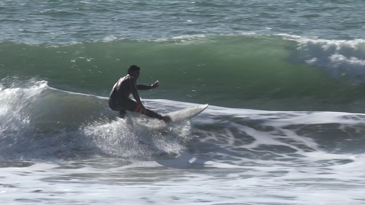 A surfer in a dark wetsuit rides low through white foam at Huntington Beach, California, with a green wave rising behind him.