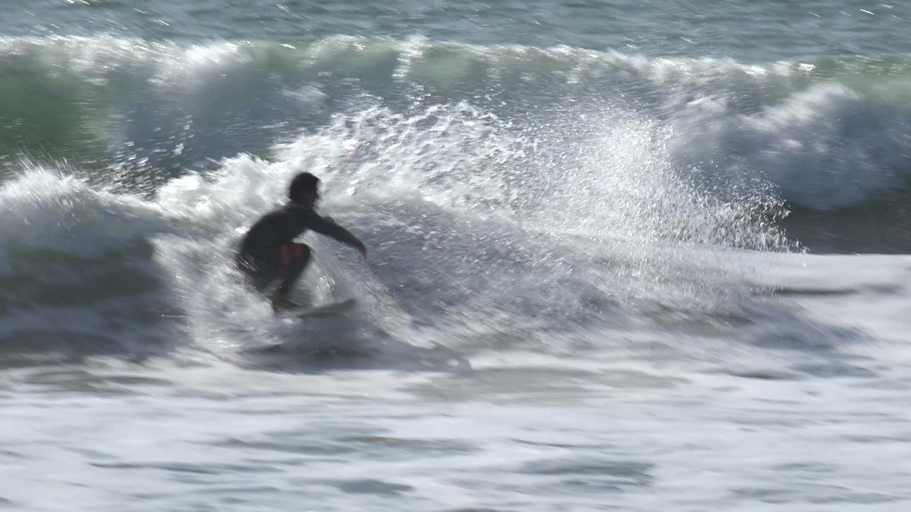 A surfer cuts across churning whitewater at Huntington Beach, California, with bright spray arcing from the breaking wave behind him.