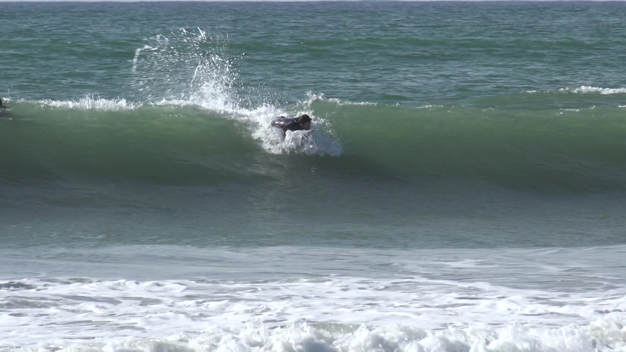 A distant surfer in a dark wetsuit rides low on a green wave at Huntington Beach, California, with white spray rising behind him and no recognizable face visible.