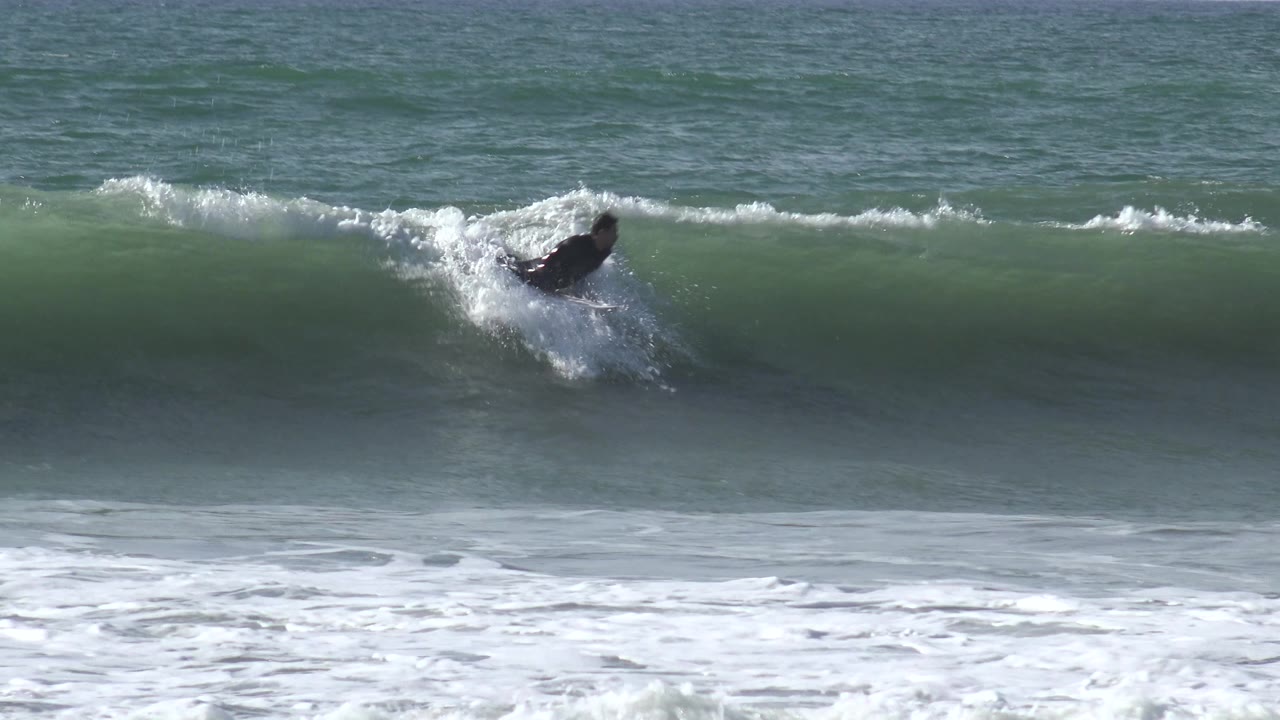 A surfer in a dark wetsuit appears on the right side of the frame riding along a curling green wave at Huntington Beach, California, with whitewater trailing behind.