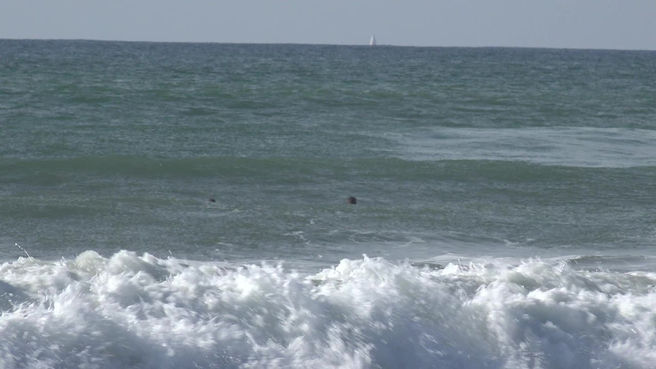 White surf rises across the lower frame at Huntington Beach, California, while small dark figures remain beyond the break under a flat ocean horizon.