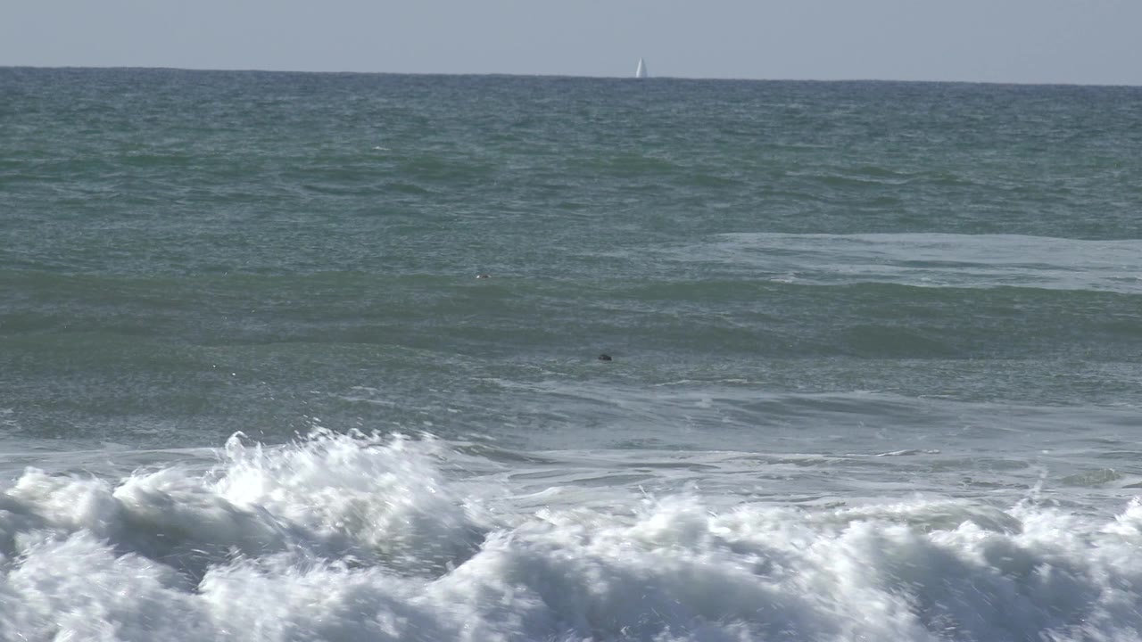 A foamy crest dominates the foreground at Huntington Beach, California, with layered ocean water, tiny offshore figures, and a small sailboat near the horizon.