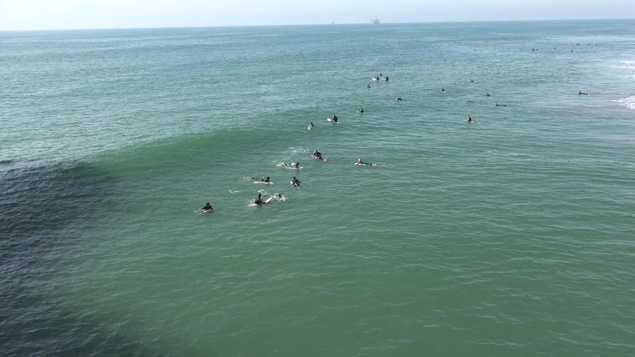 Wide Huntington Beach ocean frame with scattered surfers on boards near a low green wave and a pale horizon in bright daylight.