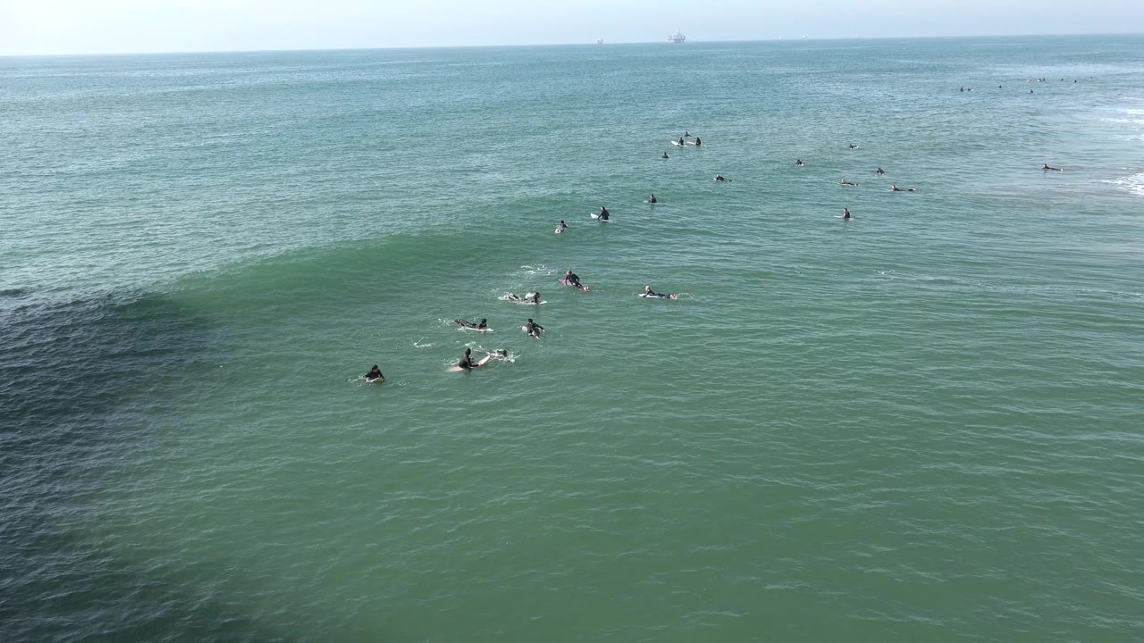 Huntington Beach surf lineup with several distant surfers paddling as a small wave begins to lift across the midground.