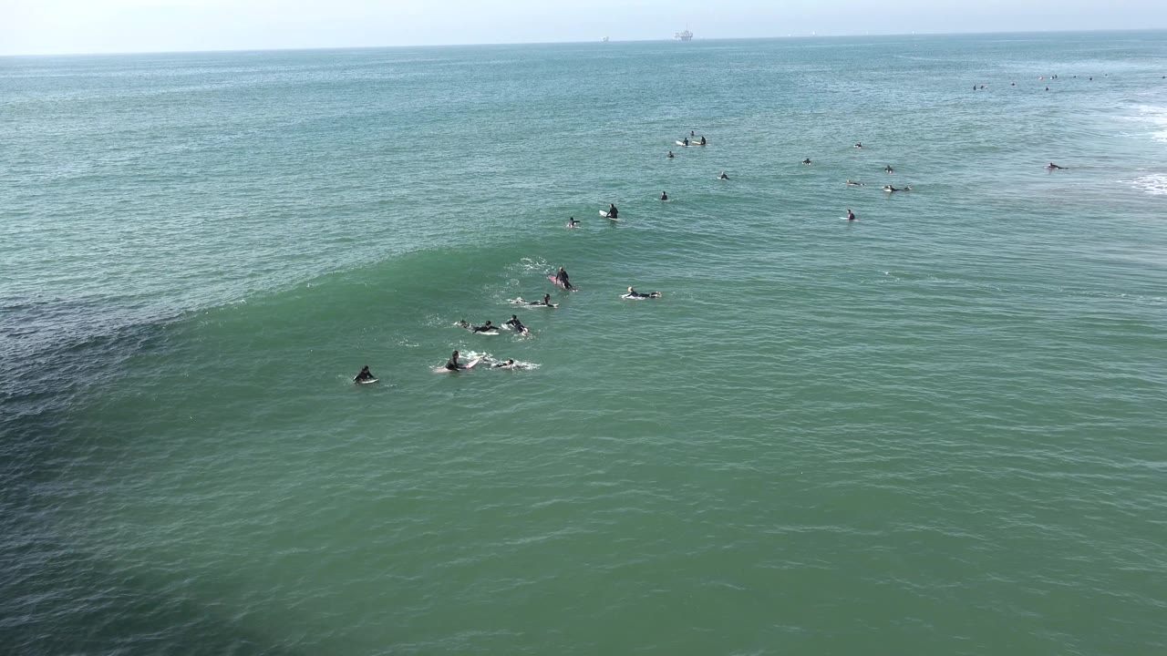 Small green wave at Huntington Beach with surfers clustered beside the break and faint whitewater farther across the ocean surface.