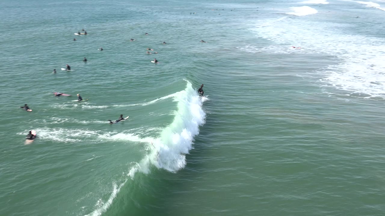 At Huntington Beach, a surfer rides along the crest of a small green-blue wave while distant surfers wait across the open water and white foam trails behind the break.