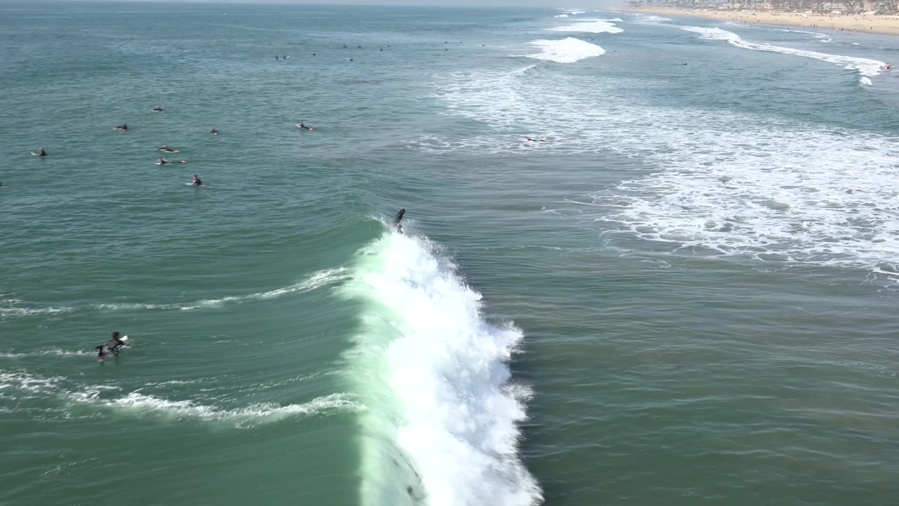 At Huntington Beach, a breaking wave throws a tall section of white spray in the midground while small surfer figures remain visible beyond the foam and the beach sits far back.