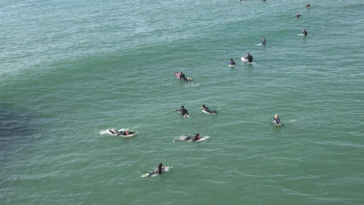 Wide view of surfers sitting and paddling across green water at Huntington Beach, California, with small ripples and sunlit surface texture.