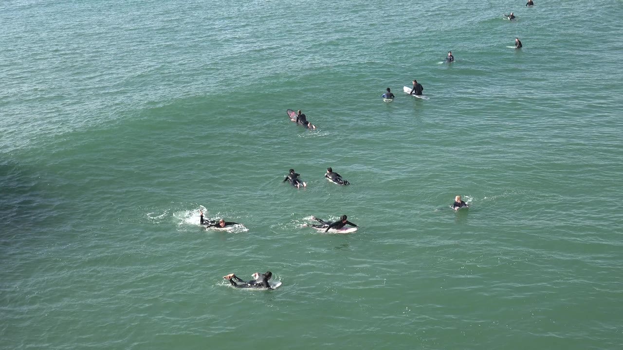 Several surfers paddle in different directions at Huntington Beach as a low green swell starts to rise across the right side of the frame.