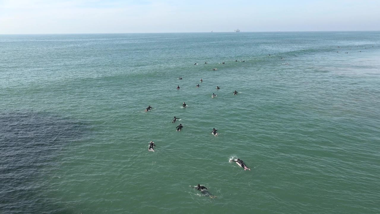 Wide view of surfers on boards spread across the ocean at Huntington Beach, with a dark patch of water in the foreground and a pale horizon beyond them.