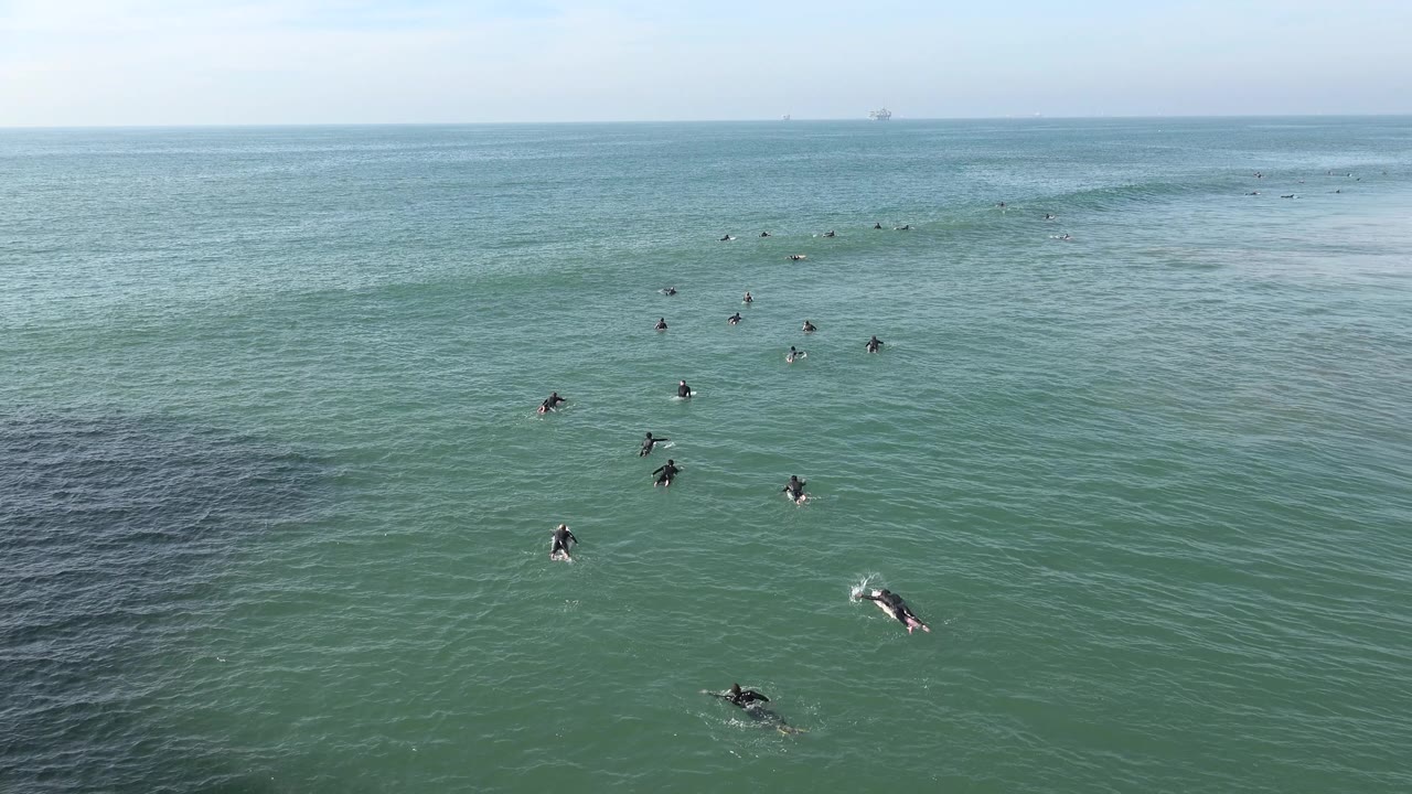 Distant surfers paddle and wait in the Huntington Beach lineup as a soft swell line stretches across the upper part of the frame.