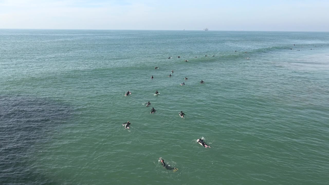 A wide Huntington Beach ocean frame shows scattered surfers below a smooth wave line, with the darker foreground water patch shifting to the left side of view.