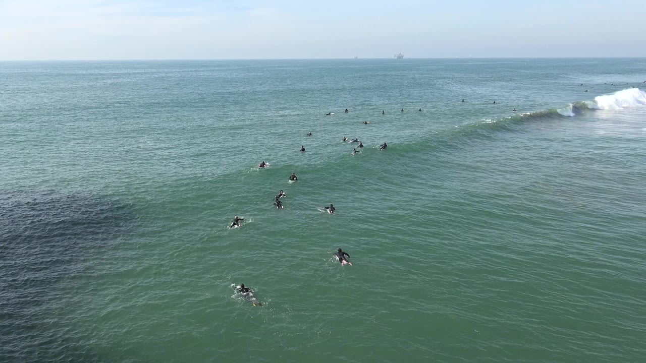 Wide Huntington Beach ocean frame with small surfers and swimmers scattered across green-blue water, a breaking wave on the right, and a pale horizon in the distance.