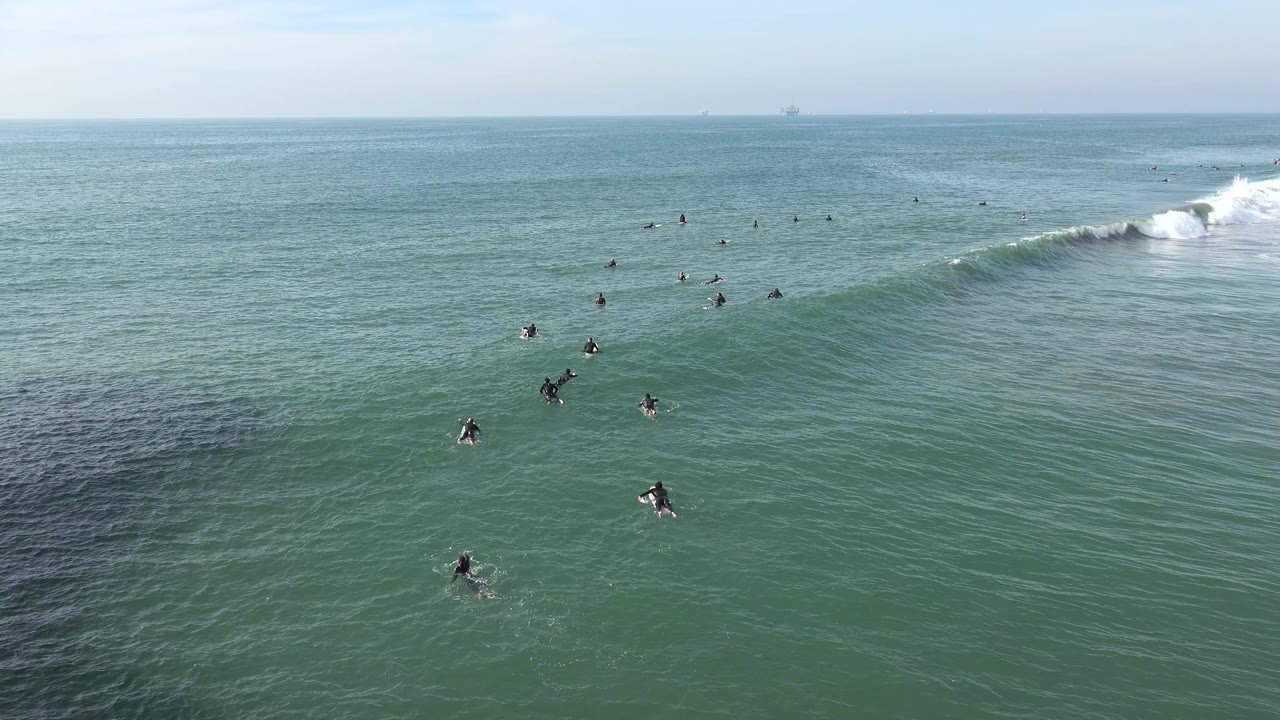 Wide Huntington Beach surf lineup with scattered people on boards, a darker patch of water in the foreground, and a white wave curling at the far right edge.