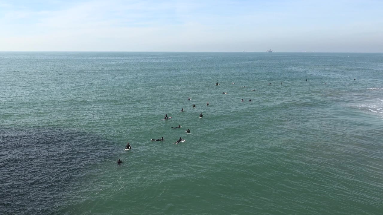 At 0:01, surfers remain spread across the Huntington Beach water while a white patch of breaking wave forms to the right of the lineup.