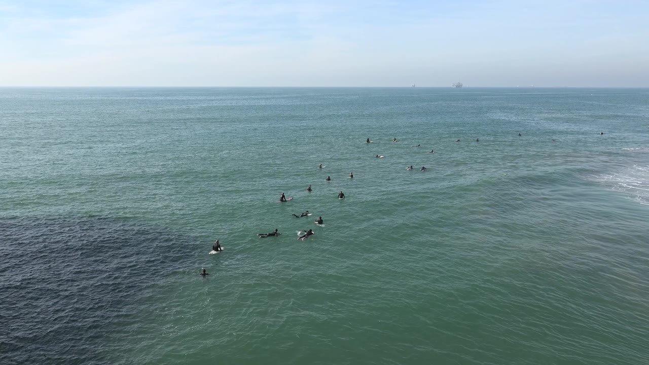 At 0:02, the Huntington Beach surf lineup appears slightly farther across the frame, with dark surfer silhouettes on boards and a low white wave edge at right.