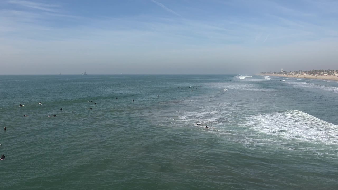 Huntington Beach waterline seen from a long distance, with small dark figures spread across the ocean, a bright white surf break, and beach visible in the right background.