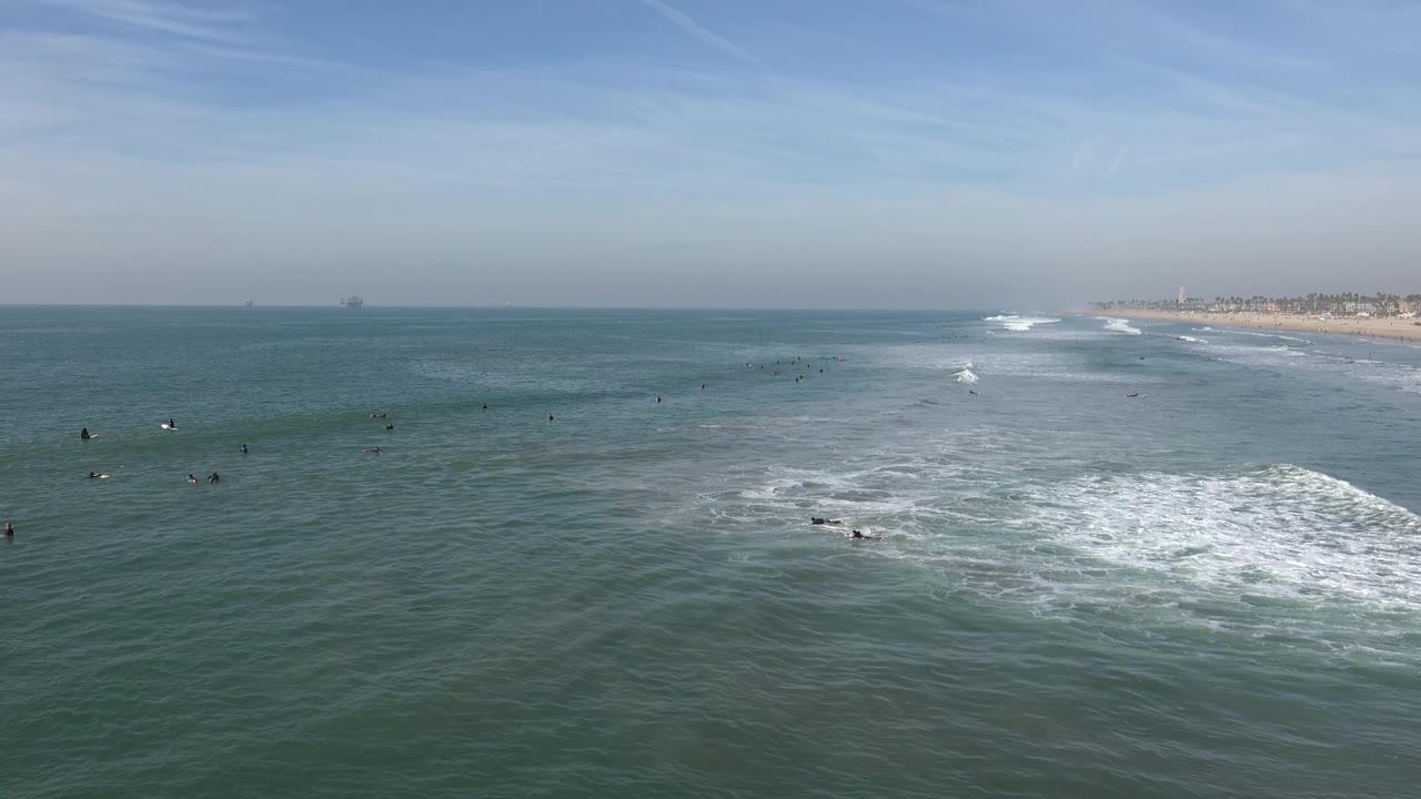Wide Huntington Beach coastal frame with scattered distant water users, rolling white surf in the midground, open sea to the left, and the sandy beach line at right.