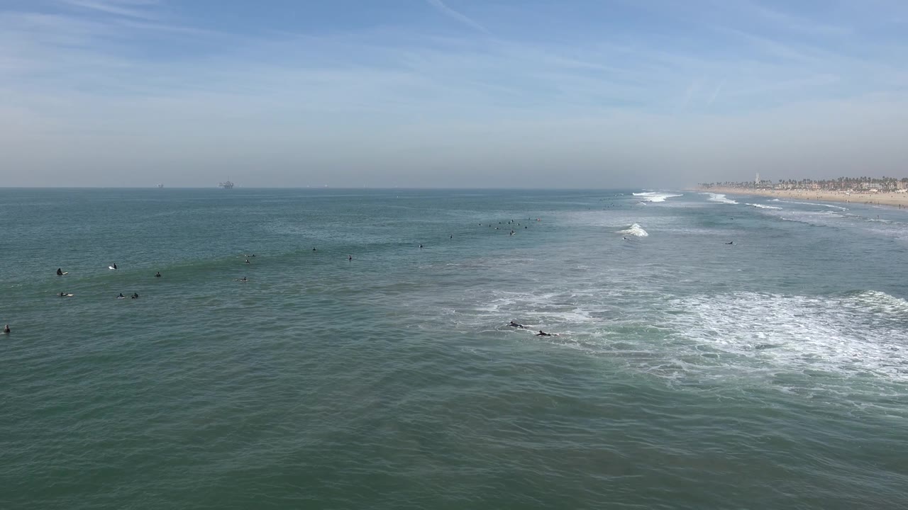 Huntington Beach ocean view with a low field of green-blue water, white surf breaking near the lower right, tiny swimmers farther out, and shoreline receding into the background.