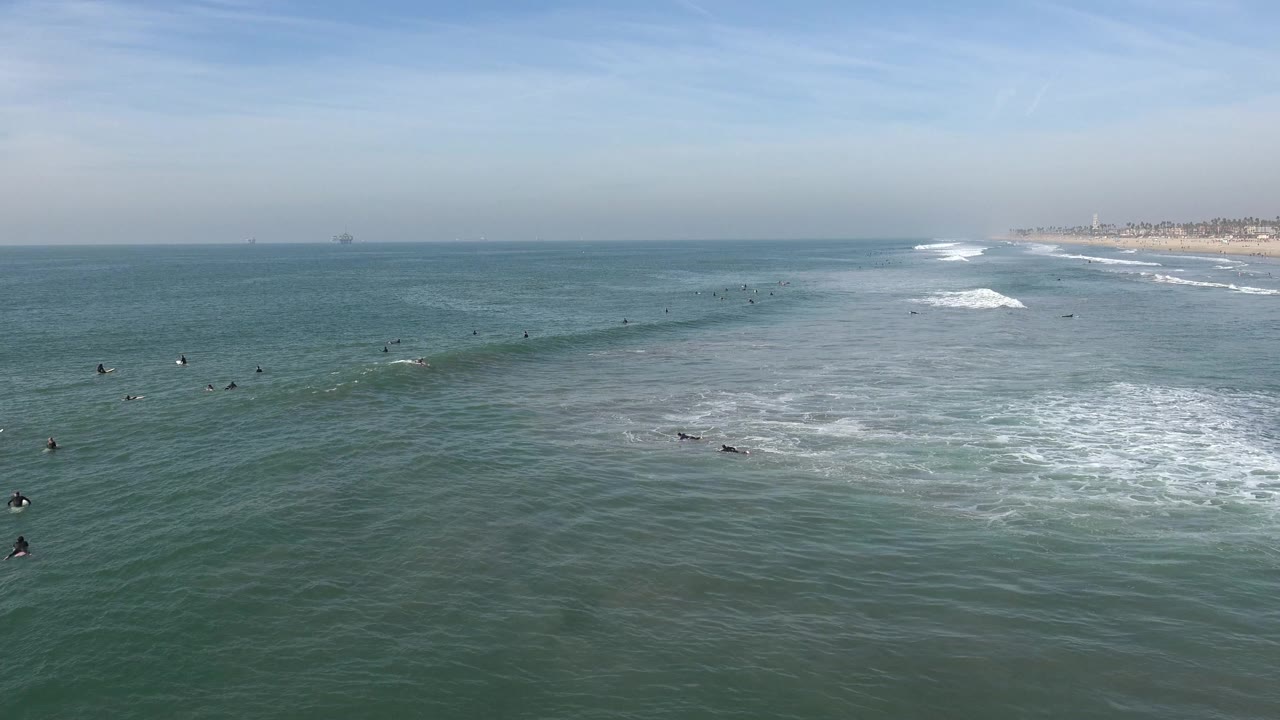 Wide view over Huntington Beach ocean water with scattered surfers and swimmers, low white surf, a pale blue sky, and the shoreline receding at the right.
