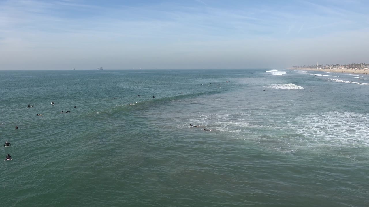 Elevated Huntington Beach view with surfers and swimmers spread across the midground, whitewash near the right side, and a faint offshore structure on the far left horizon.
