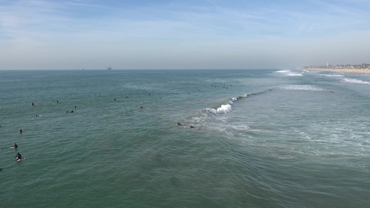 Wide view of Huntington Beach ocean water with scattered surfers and swimmers, a diagonal white wave line, hazy sky, and distant beach visible at the right edge.