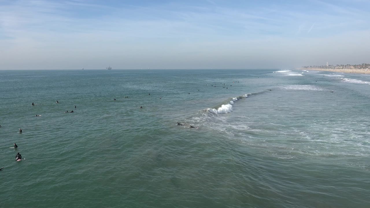 Huntington Beach surf lineup with small visible people across the midground, white water spreading from a breaking wave, and the sandy shoreline faintly visible in the background.
