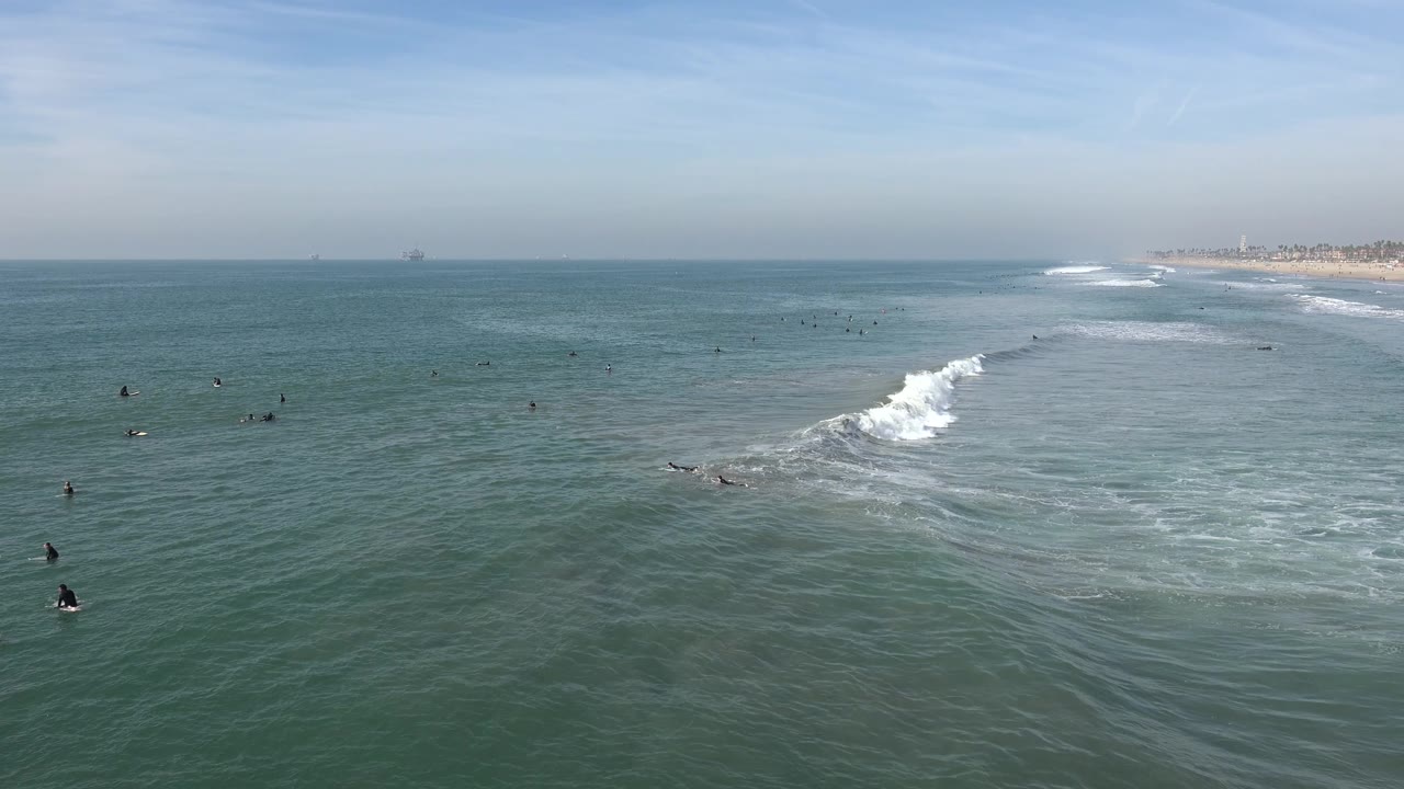Wide Huntington Beach water view with a bright breaking wave on the right, multiple surfers and swimmers waiting in the surrounding water, and a hazy coast along the far background.