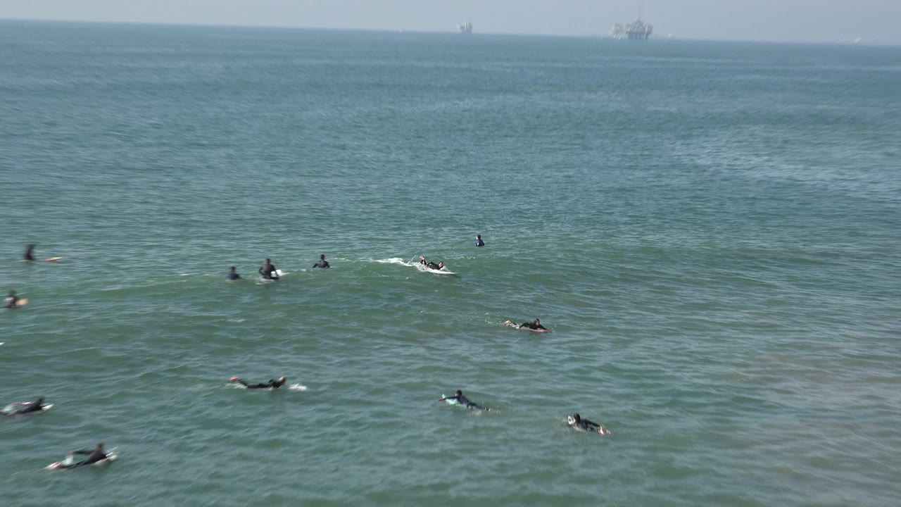 Wide frame of Huntington Beach ocean water with several distant surfers in dark wetsuits scattered across the lineup and faint offshore structures on the horizon.