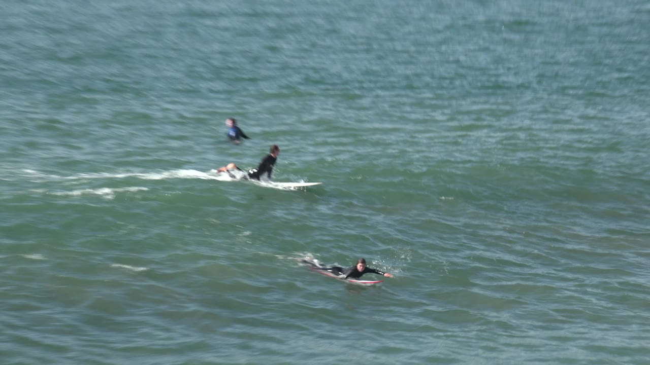 Closer Huntington Beach water view with one surfer seated on a white board, another paddling nearby, and textured green ocean filling the frame.