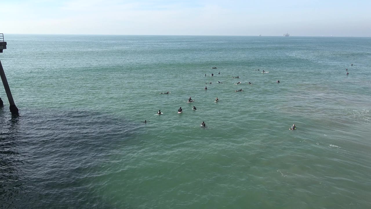 Wide Huntington Beach ocean frame with blue-green water, scattered distant figures in the surf, a pale horizon, and no recognizable people visible.