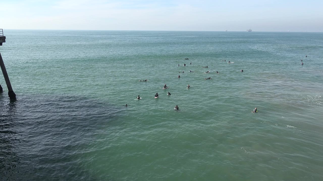 Huntington Beach water view with a left-edge vertical structure, gentle surface lines, small midground figures, and no identifiable faces visible.