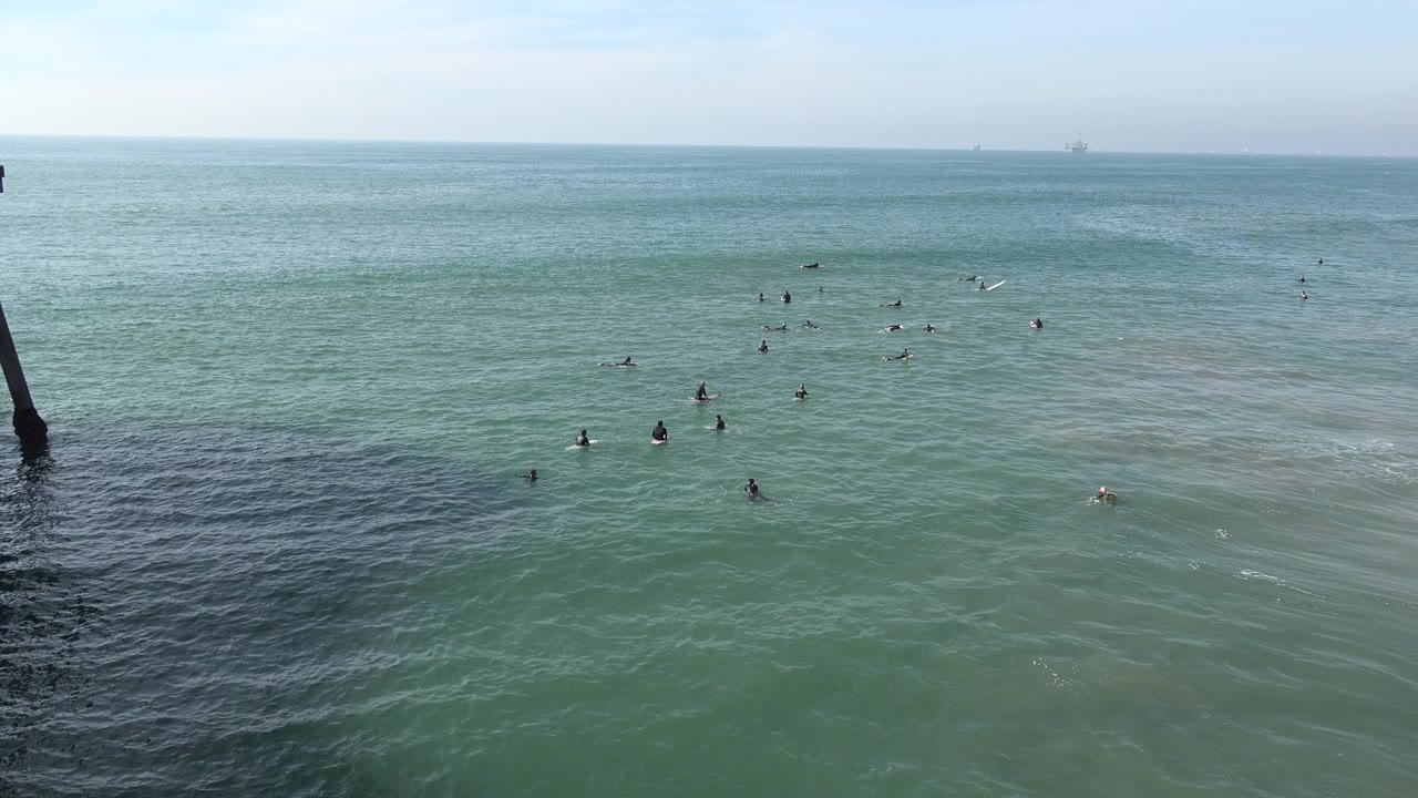 Wide Huntington Beach frame showing clustered distant figures in blue-green sea water, faint offshore structures near the horizon, and no recognizable people visible.