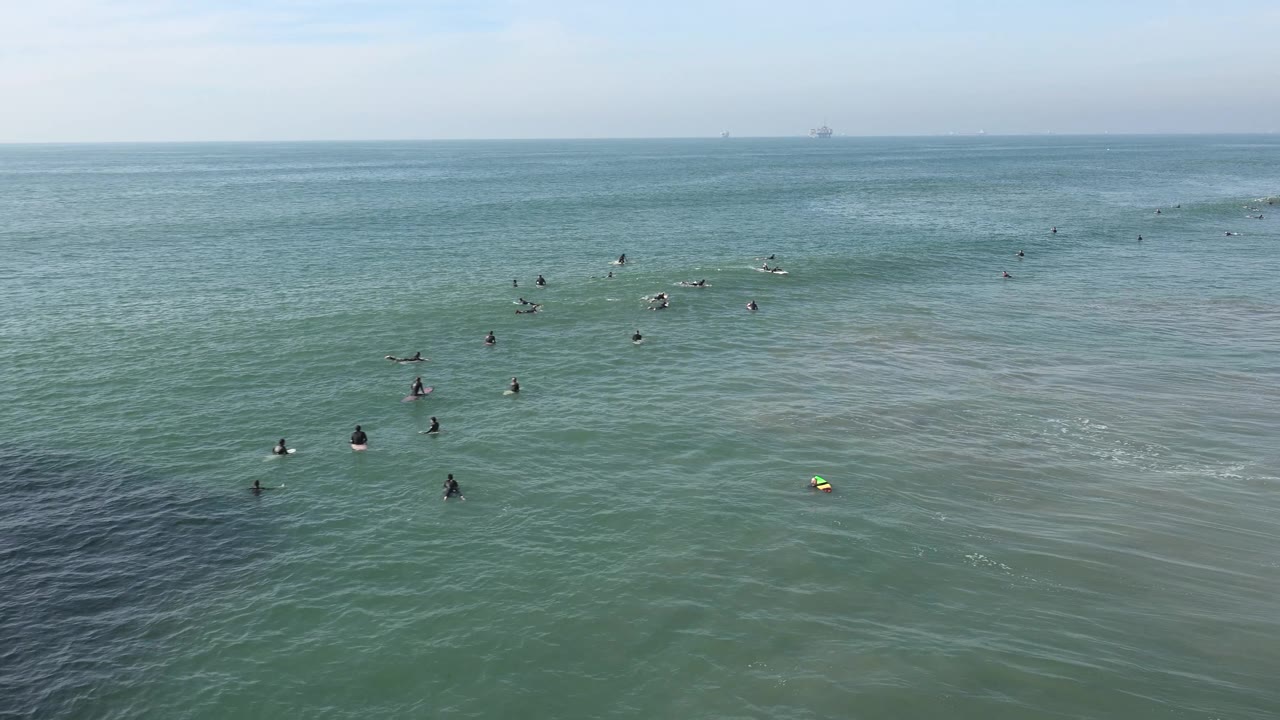 Huntington Beach surf lineup with dark-suited surfers dispersed through the midground while a broad band of textured green water fills the foreground.