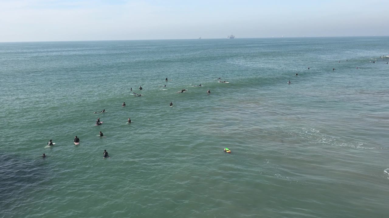 Wide Huntington Beach view with a darker swell line moving through the center of the frame and several surfers positioned around it in the distance.