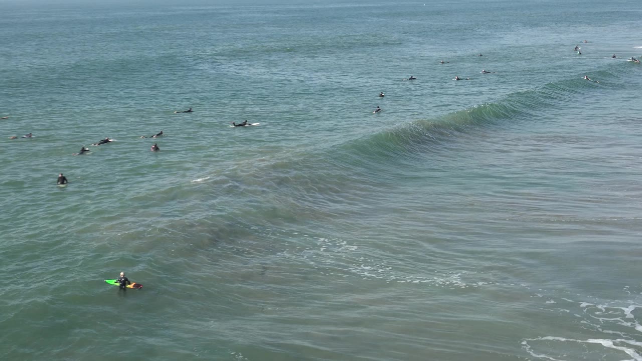 Wide Huntington Beach ocean view with scattered surfers, a green surfboard near the lower left, and a smooth swell beginning to form across the midground.