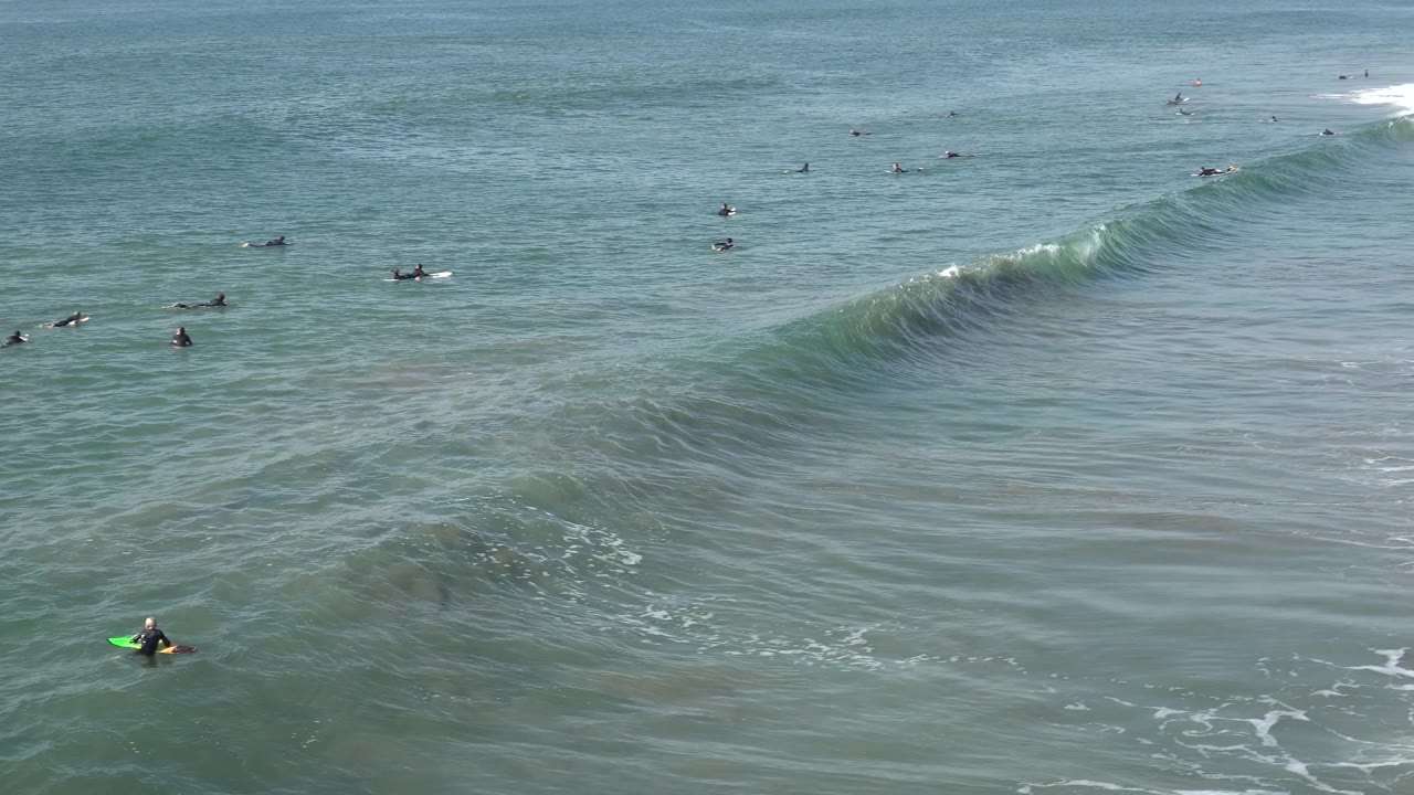 The Huntington Beach wave steepens on the right side of the frame while surfers dot the background and a near surfer with a green board stays low in the foreground water.