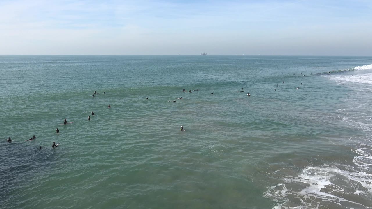Wide Huntington Beach ocean view with scattered surfers in the midground, white foam near the lower edge, and a pale horizon with distant offshore structures.