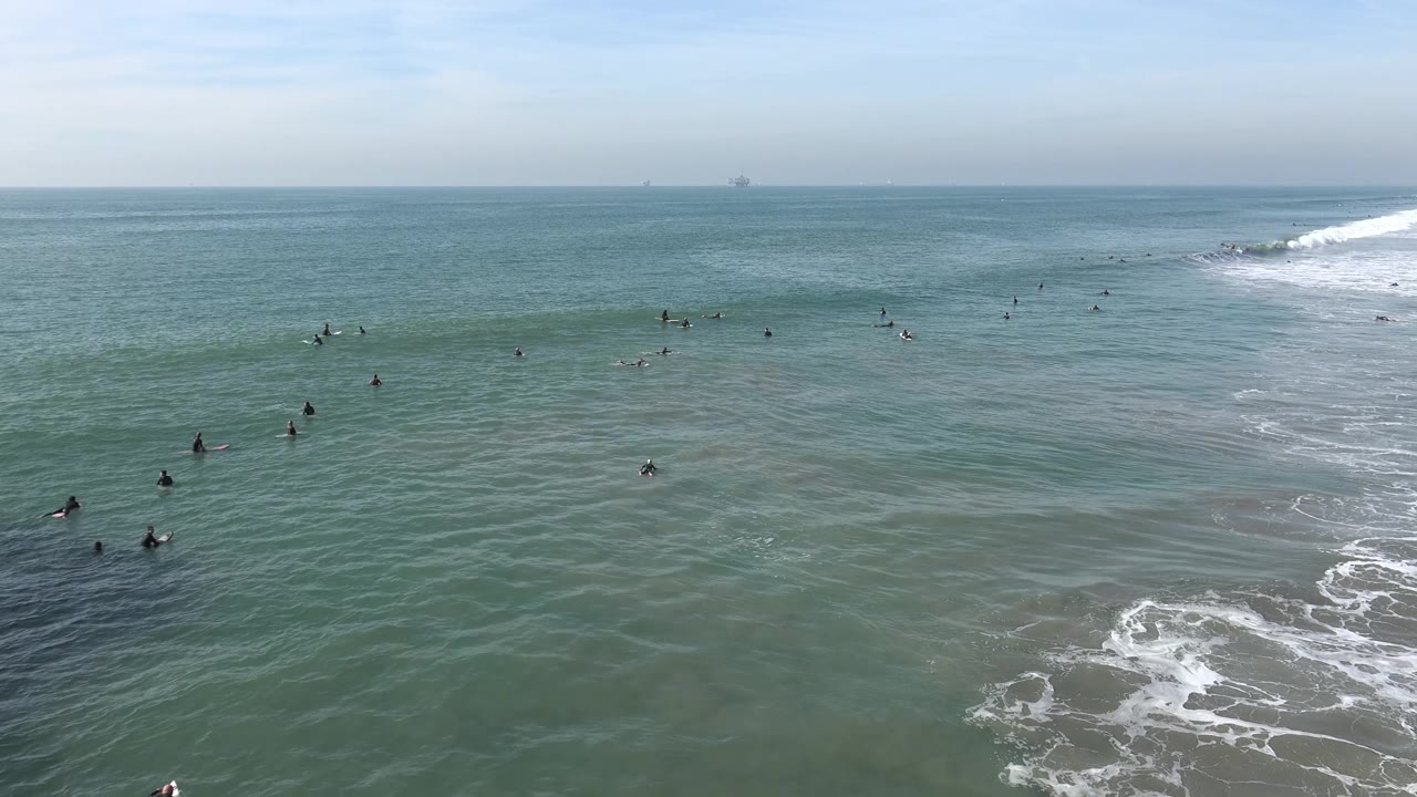 A wide Huntington Beach frame shows many small surfers in the midground, textured green water in front, and a broad whitewater band curling along the right edge.