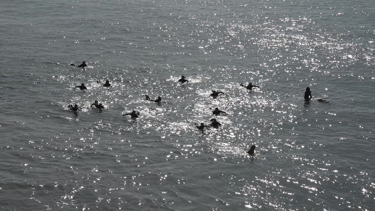 At Huntington Beach, California, distant swimmers and surfers sit across rippled water while bright sun reflections cluster near the center of the frame.