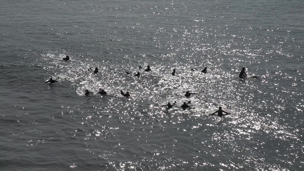 Several distant swimmers float and paddle through choppy reflective water, with no shoreline or buildings visible in the Huntington Beach frame.