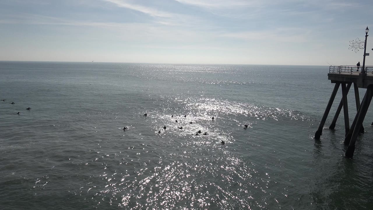 Wide Huntington Beach ocean frame with bright sun reflection on gray-blue water, small dark shapes near the surface, a level horizon, and no visible people.