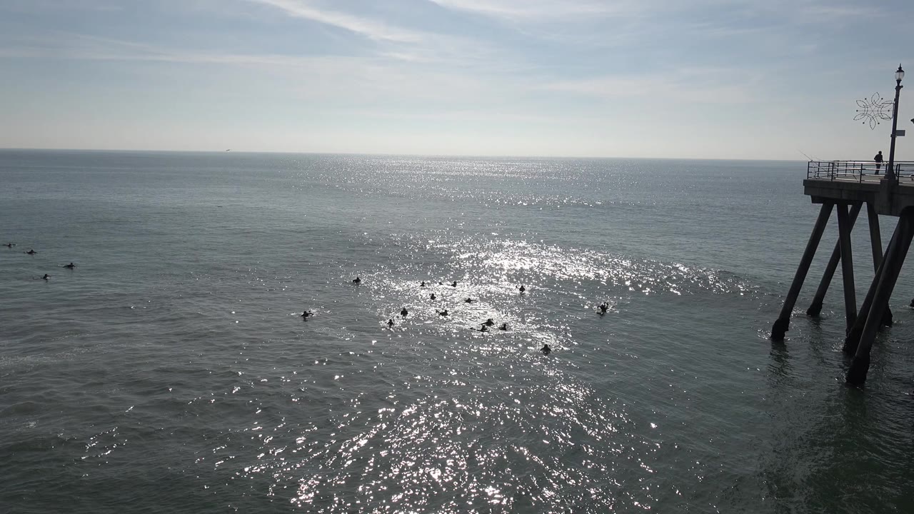 Huntington Beach water fills the frame with the strongest reflection slightly left of center, scattered dark shapes on the surface, the pier structure at right, and no visible people.