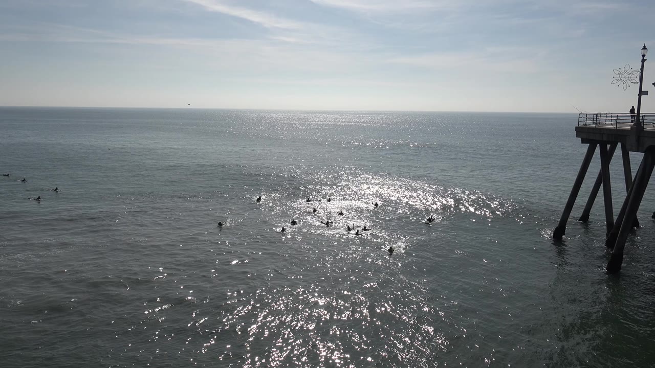 A wide Huntington Beach ocean view shows silver glare across the midground water, faint distant surface shapes, a cropped pier structure on the right edge, and no visible people.